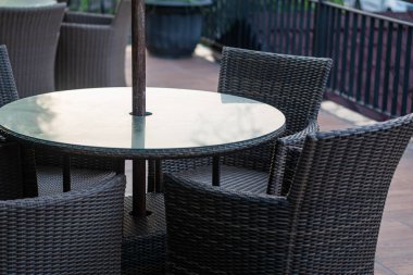 Bahçede dört hasır sandalyeyle çevrili yuvarlak cam tepeli bir masa. Dekor, yeşilliğin bulanık arka planıyla açık havada..