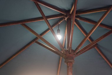 Ahşap bir şemsiye yapısının altında asılı bir ışıkla yakın çekim görüntüsü. Tahta kirişler mavi bir gölgeliği destekleyen merkezi bir kutuptan yayılıyor..