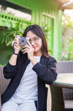 Gözlüklü genç bir kadın dışarıda bir masada oturuyor, elinde kamerayla gülümsüyor. Arkaplanda yeşil duvarlar ve bitkiler var, canlı bir atmosfer yaratıyorlar..