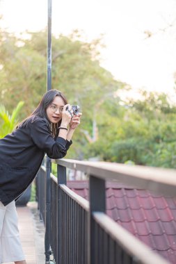 Genç bir kadın elinde kamerayla balkonda duruyor ve manzaraya bakıyor. Arkaplanda yemyeşil bir ortam ve sıcak bir günbatımı parlaklığı var..