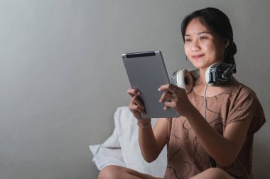 Genç bir kadın yatakta oturuyor, tablet kullanırken gülümsüyor. Boynunda kulaklık var ve rahat giyinmiş. Arka plan hafifçe aydınlatılmış, rahat bir atmosfer yaratıyor..