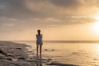 Gün batımında sakin bir sahil sahnesinde suyun kenarında durup ufka bakan bir kadın. Gökyüzü yumuşak bulutlarla ve suya yansıyan altın sarısı renklerle dolu..