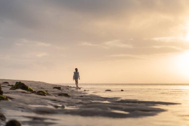 Gün batımında sakin bir sahil sahnesinde suyun kenarında durup ufka bakan bir kadın. Gökyüzü yumuşak bulutlarla ve suya yansıyan altın sarısı renklerle dolu..
