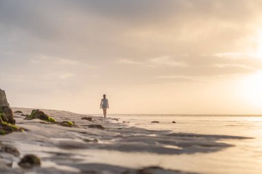 Gün batımında sakin bir sahil sahnesinde suyun kenarında durup ufka bakan bir kadın. Gökyüzü yumuşak bulutlarla ve suya yansıyan altın sarısı renklerle dolu..