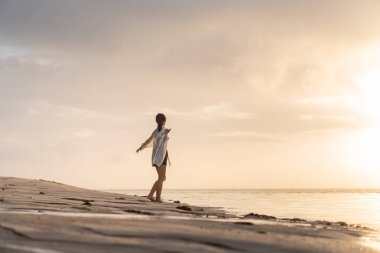 Gün batımında sakin bir sahil sahnesinde suyun kenarında durup ufka bakan bir kadın. Gökyüzü yumuşak bulutlarla ve suya yansıyan altın sarısı renklerle dolu..