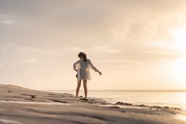 Gün batımında sakin bir sahil sahnesinde suyun kenarında durup ufka bakan bir kadın. Gökyüzü yumuşak bulutlarla ve suya yansıyan altın sarısı renklerle dolu..