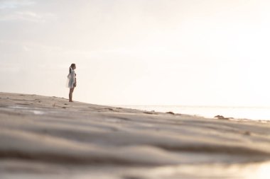 Gün batımında sakin bir sahil sahnesinde suyun kenarında durup ufka bakan bir kadın. Gökyüzü yumuşak bulutlarla ve suya yansıyan altın sarısı renklerle dolu..