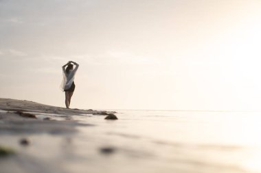 Gün batımında sakin bir sahil sahnesinde suyun kenarında durup ufka bakan bir kadın. Gökyüzü yumuşak bulutlarla ve suya yansıyan altın sarısı renklerle dolu..