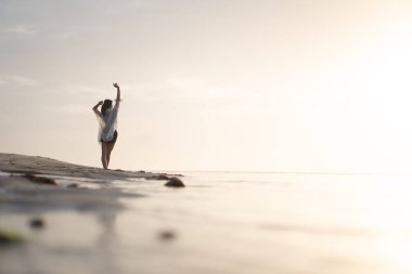 Gün batımında sakin bir sahil sahnesinde suyun kenarında durup ufka bakan bir kadın. Gökyüzü yumuşak bulutlarla ve suya yansıyan altın sarısı renklerle dolu..
