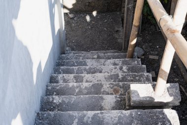 Aşağı doğru inen beton bir merdiven seti, kısmen toz ve kir ile kaplı. Merdivenler bir tarafındaki bambu korkuluklarla çevrili ve etrafındaki alan kayalık ve asfaltsız. Güneş ışığı vuruyor.