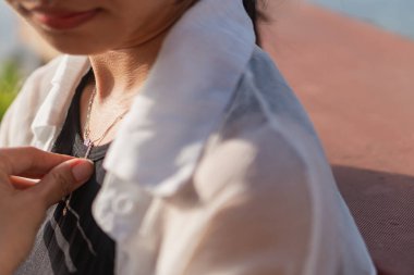 Hassas bir kolye takarken yakasını düzelten bir kadının yakın çekimi. Dikkatler boynuna ve mücevherlerine çevrildi. Yumuşak doğal ışıklandırmayla özellikleri vurgulanıyor. Arkaplan bulanık,