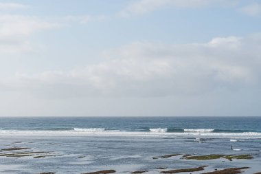 Parçalı bulutlu bir gökyüzünün altında kıyıya vuran yumuşak dalgaların olduğu sakin bir kıyı sahnesi. Ufuk sakin okyanusa uzanırken ön planda ıslak kum ve kayalık bölgeler görülüyor..