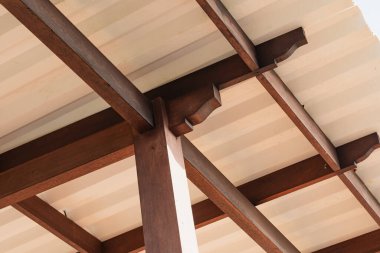 Açıkta duran kirişler ve üzerinde tek bir ampul olan ahşap tavan yapısının yakın çekim görüntüsü. Tavanda ahşap ve metal elementlerin birleşimi modern bir estetik yaratıyor..