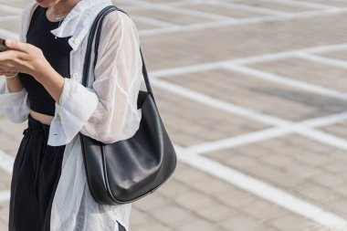 Boş bir otoparkta tek başına duran genç bir kadın, siyah bir bluz ve şort giyiyor, açık renk bir hırkayla. O, arka planda park etmiş bir motosikletle akıllı telefonuna odaklanmış..