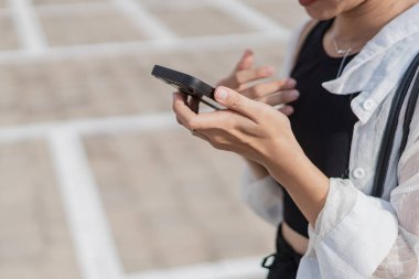 Boş bir otoparkta tek başına duran genç bir kadın, siyah bir bluz ve şort giyiyor, açık renk bir hırkayla. O, arka planda park etmiş bir motosikletle akıllı telefonuna odaklanmış..
