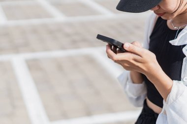 Boş bir otoparkta tek başına duran genç bir kadın, siyah bir bluz ve şort giyiyor, açık renk bir hırkayla. O, arka planda park etmiş bir motosikletle akıllı telefonuna odaklanmış..