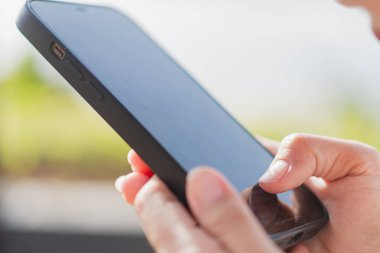 Akıllı bir telefon tutan ve ekrana odaklanan birinin yakın çekimi. Arka plan bulanık, yeşillik ve açık hava manzarası var. Adamın parmakları nazikçe ekrana dokunuyor.