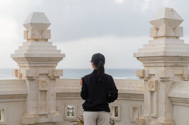 Siyah ceketli ve şapkalı bir kadın, bulutlu bir gökyüzünün altında okyanusa bakarken elinde kırmızı bir kahve fincanı tutan bir taş korkuluğun yanında duruyor..