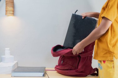 Bir öğrenci okula hazırlanmak için büyük bir kitabı kırmızı bir sırt çantasına koyar. Arkada rahat bir lamba ve istiflenmiş kitaplar var..