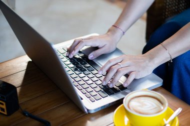 Bir insanın ellerinin dizüstü bilgisayarda klavyeye yazışının yakın çekimi. Sahnede bir ahşap masa, bir fincan kahve fincanı ve bir fincan kahve tabağı ve küçük bir kamera yer alıyor. Şey...