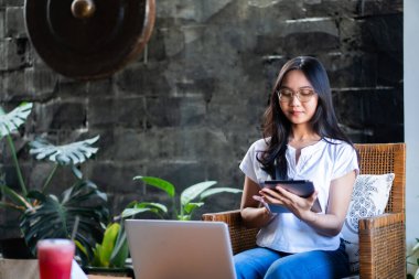 Kapalı bir ortamda rahatça oturup tablet kullanan genç bir kadın. Uzun saçları var ve gözlük takıyor. Sıradan beyaz bir bluz ve kot pantolon giyiyor. Öne doğru.