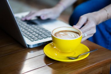 Sarı kahve fincanının yakın plan resmi, bir fincan tabağında latte resmi, ahşap bir masanın üzerine konmuş. Bir kişi arka planda dizüstü bilgisayarda yazı yazıyor, samimi bir çalışma ya da çalışma öneriyor.