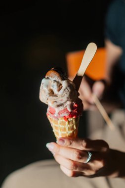 İki kepçeli dondurma külahını tutan bir elin yakın çekimi: bir çikolata ve bir çilek. Koni klasik bir waffle stilidir ve içine tahta bir çubuk yerleştirilir.