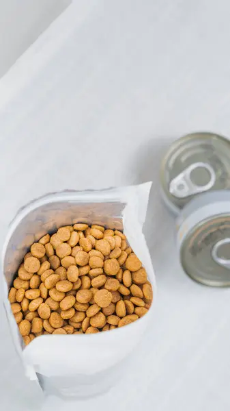 stock image A top view of a partially opened bag of pet food, showcasing the kibble inside. Next to the bag, there are two cans, possibly of wet pet food, with pull-tab lids. The cab