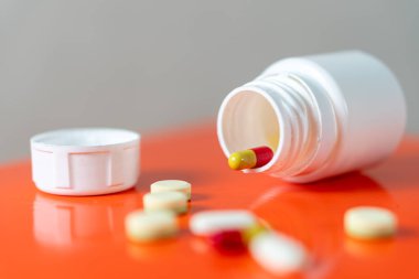 Kapağı açık beyaz bir hap şişesi, yan yatmış, çeşitli haplar ve kapsüller turuncu bir yüzeye dökülmüş..