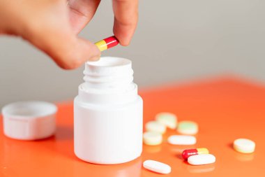 Kırmızı ve sarı kapsülü turuncu bir yüzeye yayılmış çeşitli haplarla beyaz bir ilaç şişesinin üzerinde tutan bir el..
