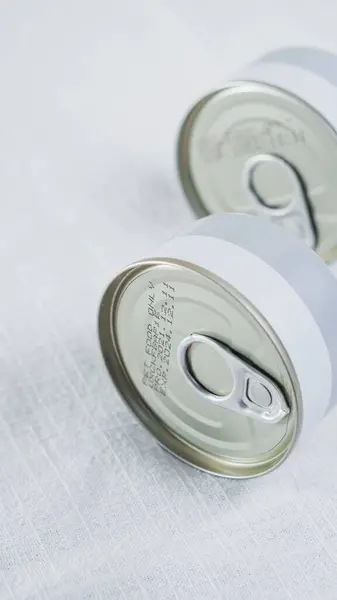 Açık dokulu bir yüzeye yerleştirilmiş, çekiş-tab kapaklı iki alüminyum kutu. Kutuların ortasında beyaz bir şerit olan minimalist bir tasarımı var.