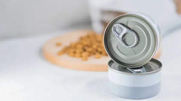 Beyaz bir masa örtüsünün üzerinde iki adet metal kutu ve arka planda ahşap kesim tahtası ve evcil hayvan yiyeceği var. Kutular çekiş kapaklı gümüş, bu da F içerdiğini gösteriyor.