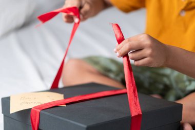 Bir çocuk Noel hediyesini açıyor ve kırmızı kurdelelere odaklanıyor. Hediye kutusu siyah ve üzerinde Mutlu Noeller yazan bir etiket var. Çocuk bir yatakta oturuyor, üzerinde