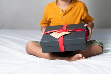 Yatakta oturan bir çocuk, elinde kırmızı kurdeleyle sarılı siyah bir hediye kutusu var. Kutuda 