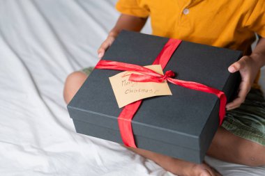 Kırmızı kurdeleli siyah bir hediye kutusu ve üzerinde mutlu Noeller yazan bir etiket tutan bir çocuk! Bir çocuk beyaz bir yatakta oturuyor, sarı bir gömlek giyiyor.