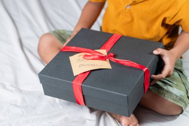 Yatakta oturan bir çocuk güzel paketlenmiş kırmızı kurdeleli siyah bir hediye kutusu tutuyor. Kutuda 