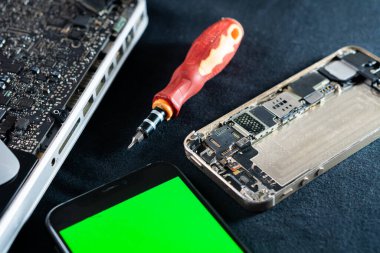 Laptopun anakartının yanında parçalanmış akıllı bir telefonun yakın çekim görüntüsü. Akıllı telefonun yanına kırmızı ve siyah bir tornavida yerleştirilir. Akıllı telefon ekranı yeşil.