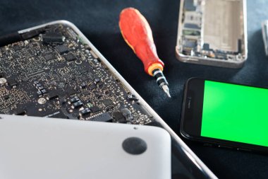 Parçalanmış elektronik cihazların yakın çekim görüntüsü. Laptop ana kartı, akıllı telefon ve tornavida da dahil. Dizüstü bilgisayarın iç parçaları görünür, vitrin.