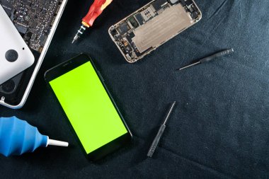 Yeşil ekranlı akıllı bir telefon siyah kumaş bir yüzeye yerleştirilir, tornavida, üfleyici ve sökülmüş bir telefon gibi çeşitli araçlarla çevrilidir. Arkaplan