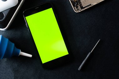 Parlak yeşil ekranlı, karanlık bir yüzeye yerleştirilmiş akıllı bir telefon. Yakınlarda mavi bir tutkal şişesi ve küçük bir tornavida var. Tamir ya da bakım seti öneriyor..
