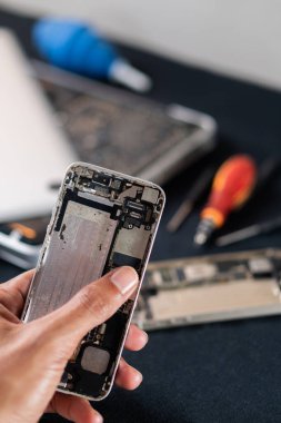 Parçalanmış akıllı bir telefonu tutan ve içindeki bileşenleri açığa çıkaran bir kişinin yakın çekimi. Arkaplan, bir onarım veya montaj çalışma alanını gösteren araçlar ve diğer elektronik aygıtlar içerir.