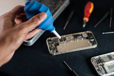 Siyah bir yüzey üzerinde parçalanmış akıllı telefonu temizlemek için mavi hava üfleyicisi kullanan biri..