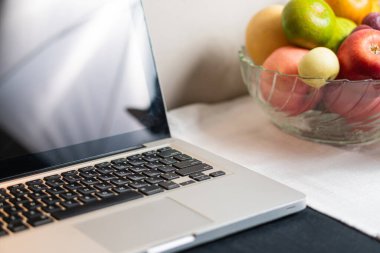 Dizüstü bilgisayarın klavyesinin yakın plan görüntüsü bir kase meyvenin yanında. Dizüstü bilgisayarda siyah ekran var. Kasede elmalar, armutlar ve diğer renkli şeyler var.