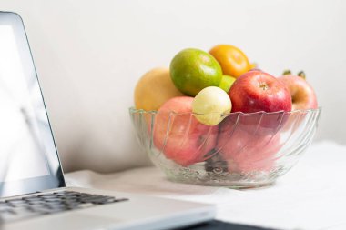 Elmalar, limonlar ve limonlar da dahil olmak üzere çeşitli taze meyvelerle dolu temiz bir cam kase beyaz bir masa örtüsünün üzerinde bir dizüstü bilgisayarın yanına yerleştirilmiş..