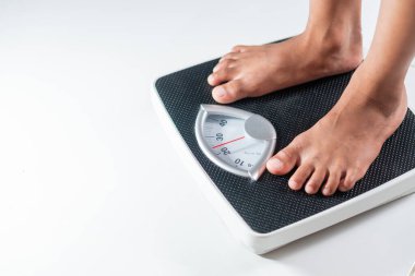 Mekanik bir banyo terazisinde duran bir insan, ayaklarının yakın çekimini ve ölçeğin kadranını gösterir. Arka plan düz beyaz, ölçeği ve ayakları vurguluyor..
