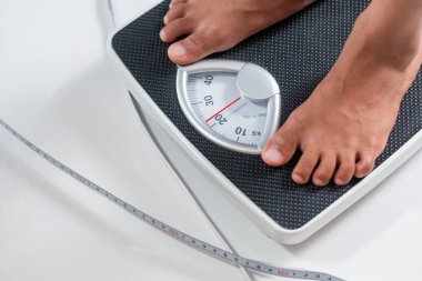 Mekanik tartı terazisinde duran birinin yakın çekimi. Ölçek 70 kg gösteriyor. Tartının yanına bir mezura yerleştiriliyor.