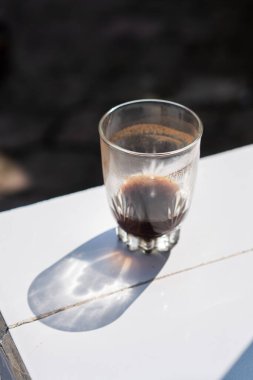 Koyu sıvılı bir cam beyaz bir yüzeye oturur ve gölge bırakır. Bardak kısmen kahve ya da çaya benzeyen bir şeyle dolu, koyu kahverengi bir rengi gösteriyor. Şey...