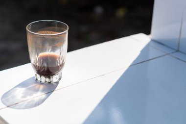 A glass of dark coffee sits on a white tiled surface, illuminated by sunlight. The glass is half full, casting a shadow on the tiles, creating a contrast between light and dark. clipart