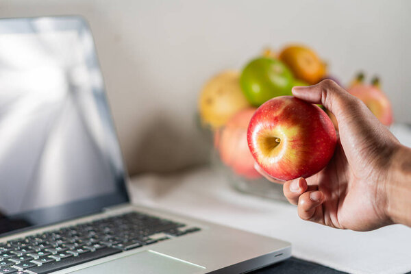 Человек, держащий красное яблоко перед ноутбуком, с миской сортированных яблок на заднем плане. Сцена хорошо освещена, демонстрируя современное рабочее пространство с акцентом на здоровое питание.