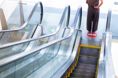 Yürüyen merdivende duran, aşağı bakan biri. Yürüyen merdivende hareketin akışını gösteren net yön işaretleri var. Dekor modern bir iç mekan gibi görünüyor.